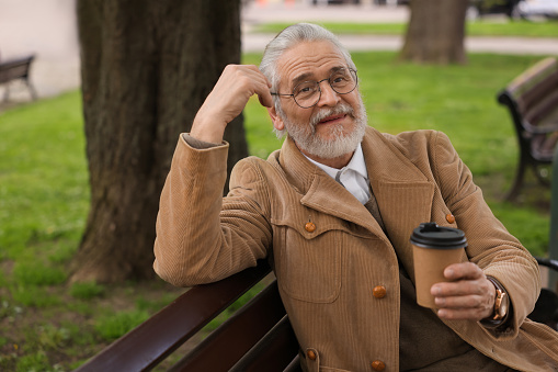 Handsome senior man sitting on bench and drinking coffee outdoors, space for text