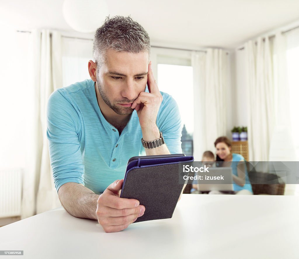 Adult Man Reading Book on E-Reader Focus on an adult male reading a book on an e-reader at home, while his wife and son sitting on sofa in the background and using laptop.  Laptop Stock Photo
