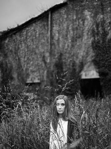 A young woman looks at the camera, in a wild place, hidden among tall grasses, near an abandoned place