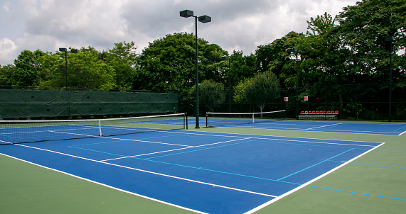 Cross court view of pickleball tennis courts painted blue and green with lights and trees in the background.