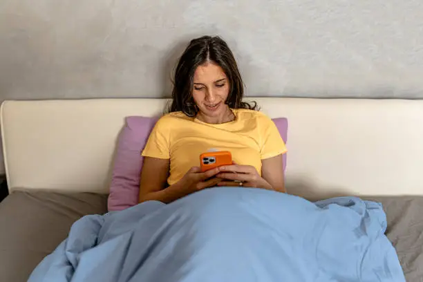 A young woman, illuminated by the glow of her phone, types away on her device while reclining in bed, presumably sending a late-night message
