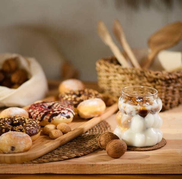 A wooden platter holds donuts, chocolate-filled cookies, and bite-sized cookies. Behind, walnuts rest on a white cloth, with wooden utensils in a wicker basket. An iced milk with espresso completes the scene