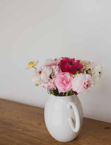 Vertical closeup of mixed ranunculus in white jug on timber table against plain background wth vintage filter effect (selective focus)