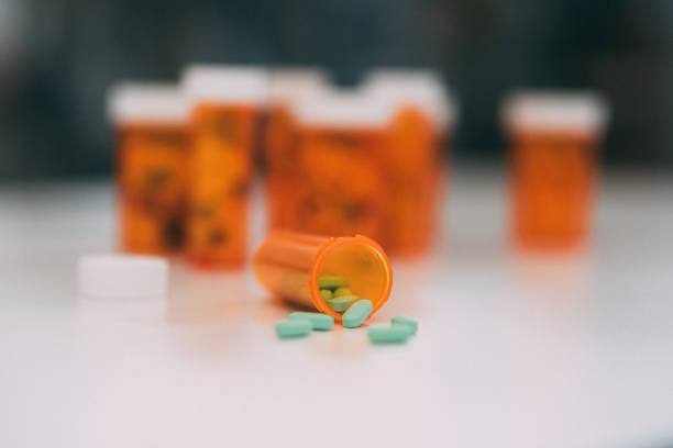 An orange medicine bottle sitting on its side with green medical pills spilling out. The cap to the bottle is sitting nearby on the countertop with many other pill bottles.