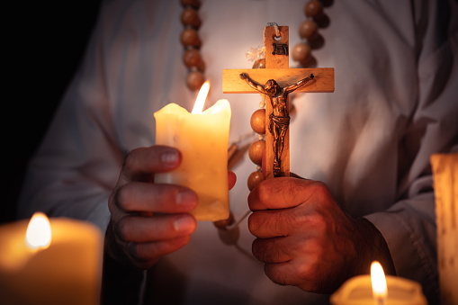 In the candlelight, the priest n a white priest's robe holds in his hands a wooden rosary with a wooden cross and Christ on it.