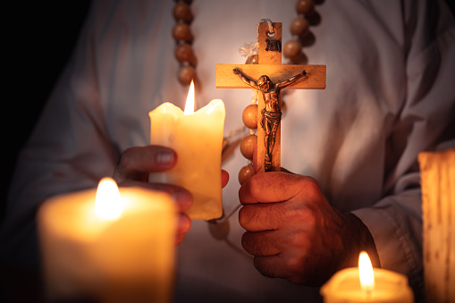 In the candlelight, the priest n a white priest's robe holds in his hands a wooden rosary with a wooden cross and Christ on it.