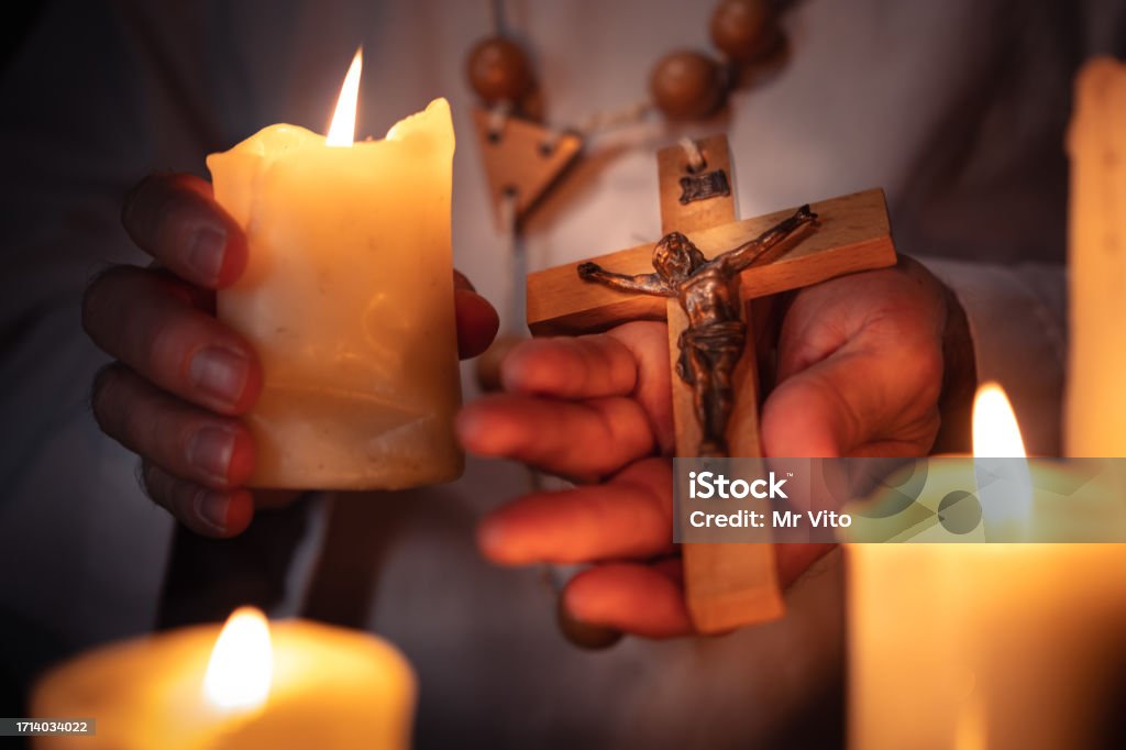 A priest with a cross in his hand In the candlelight, the priest n a white priest's robe holds in his hands a wooden rosary with a wooden cross and Christ on it. 55-59 Years Stock Photo