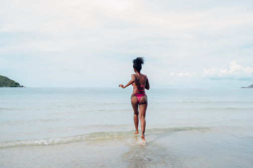Beautiful African american woman in pink bikini on tropical beach. Portrait of dark skinned woman smiling at sea. Brunette tanned girl in swimwear enjoying and walking on beach.
