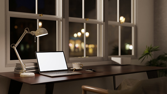 Side view image of a modern office workspace at night with a white-screen laptop mockup, a table lamp, and accessories on a table against the window. 3d render, 3d illustration