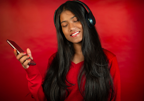 Portrait of beautiful, happy woman young woman with long black hair wearing a red sweater and listening music through headphones with holding smartphone in hand against the plain red background.