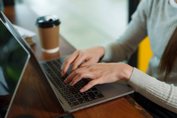 Clous-up hands of Asian businesswoman wearing long-sleeved blue sweater Typing work checking sales small business cafe laptop on wooden table brown disposable paper coffee cup. there's phone on it manos de una mujer de negocios asiática con suéter azul de manga larga trabajo de mecanografía de verificación de ventas, cafetería de pequeñas empresas, computadora portátil sobre mesa de madera, taza de café de papel marrón desechable. hay un te