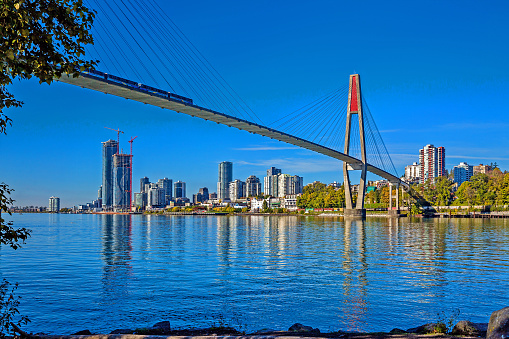 Skytrain bridge connecting Surrey and New Westminster over the Fraser River, New Westminster city center on the opposite bank, two high-rise buildings under construction, blue sky in the background