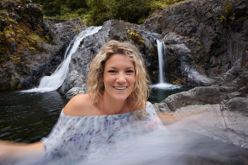 Young happy woman having fun by the waterfall in nature and looking at camera. Blurred motion.