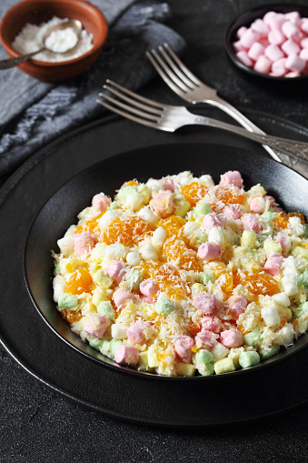 Ambrosia Salad with pineapple, mandarin oranges, yogurt, mini marshmallows, coconut and whipped cream in black bowl on concrete table, american recipe, vertical view, close-up