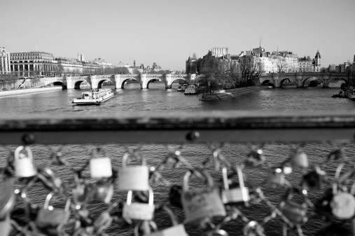 Black and white image taken from the Pont des Arts in Paris, looking towards the Ile de la Cite and Notre Dame cathedral. A 'bateau mouche' is about to pass under the Pont Neuf; out of focus in the foreground, there are love padlocks attached to the metal grill of the Pont des Arts bridge.