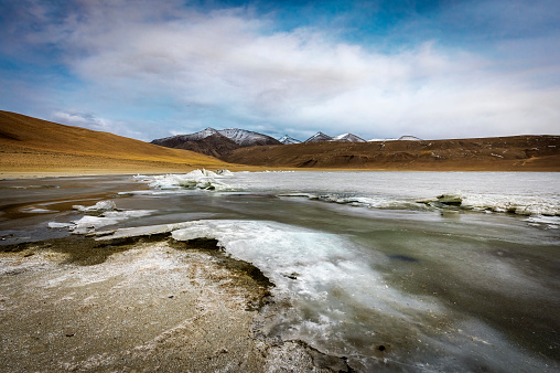 Nature's artistry on display at Kyagar Tso Lake in Ladakh, with ice formations along the shore, under an overcast sky, and distant snowcapped peaks