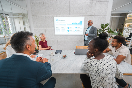 Senior mixed race businessman leads a dynamic knowledge exchange, engaging coworkers and reinforcing insights with data displayed on a screen.