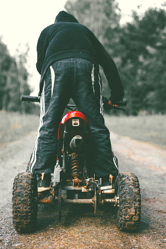 A child boy stands on an ATV wearing a black hoodie with his back to the camera. Quad bike. In the forest on a dirt road, a child stands riding his ATV. A child riding a small quad bike..