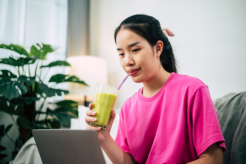 Portrait of Young sporty fit slim asian woman coach using laptop and drinking green smoothies of spinach and banana. She exudes relaxation and calmness, embodying the healthy lifestyle concept.