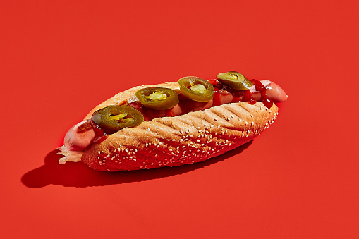 American hot dog with jalopeno on red background minimal style. Fast food on colour background with hard shadow. Sandwich with sausage trendy concept