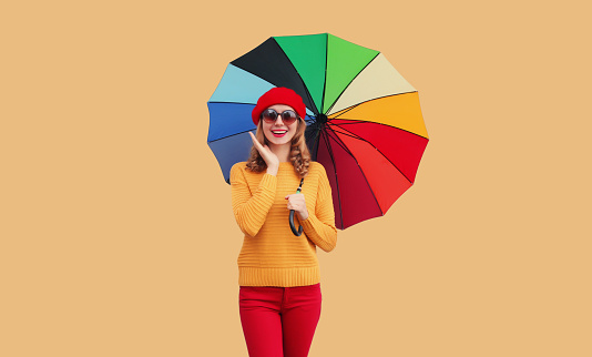 Autumn color style outfit, beautiful happy cheerful young woman holds colorful umbrella enjoys new season in french beret hat, yellow sweater on beige studio background