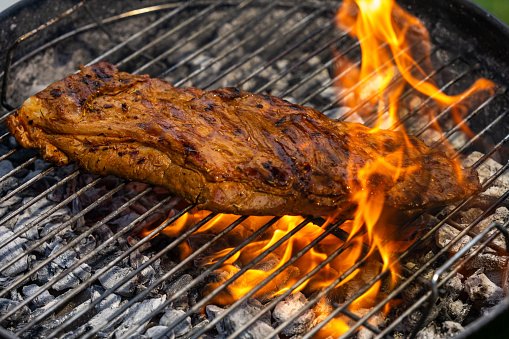 Piece of pork engulfed in flames roasting on a wire grid of a charcoal barbecue grill.