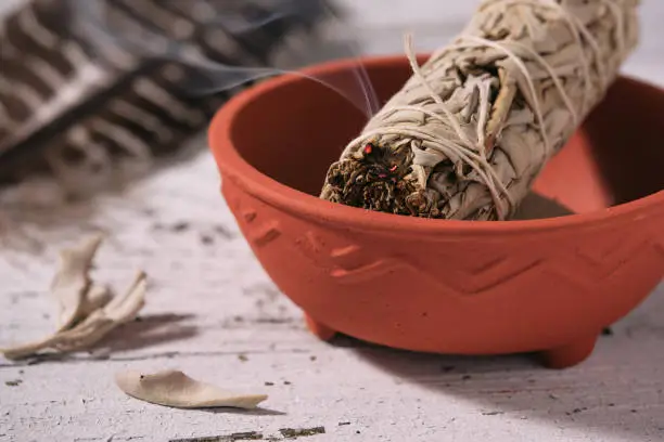 Burning White Sage smudge stick (Salvia apiana) in a clay bowl and smudging feather on an old white wooden table. White sage is traditionally used by Native American peoples for spiritual clearing and healing. High angle view, close-up, photographed on a sunny day.