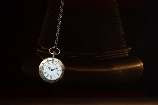 Hypnosis session. Vintage pocket watch with chain swinging over surface on dark background among faded clock faces, magic motion effect