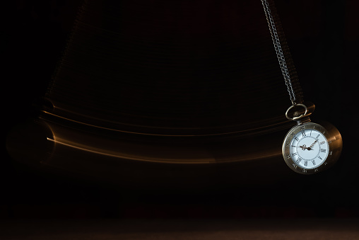 Hypnosis session. Vintage pocket watch with chain swinging over surface on dark background among faded clock faces, magic motion effect