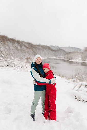 Mother embraces daughter having fun walking in snow nature near lake. Happy mom and child hugging in winter park, spending holidays. Family walk in mountain country in snowy forest together.
