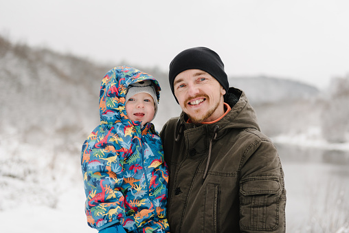 Father hugs little baby son having fun walking in snow nature together. Happy dad and kid playing in winter park, spending holidays. Family hugs in mountain country in snowy forest together. Closeup.