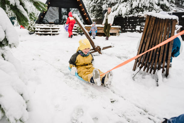 winterurlaub im freien. familie mit kindern amüsiert sich im hinterhof des berglandhauses im schneewald. mutter, tochter, sohn gehen. vater zieht kleinen sohn auf schlitten während schneefall am wintertag. - little boys sled clothing slide stock-fotos und bilder