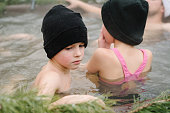 Children in hats relax in hot bath outdoors. Kids enjoying thermal spa in snowy forest. Winter holidays in mountains, hot water treatments concept. Family in hot tub open air while snowing. Closeup.