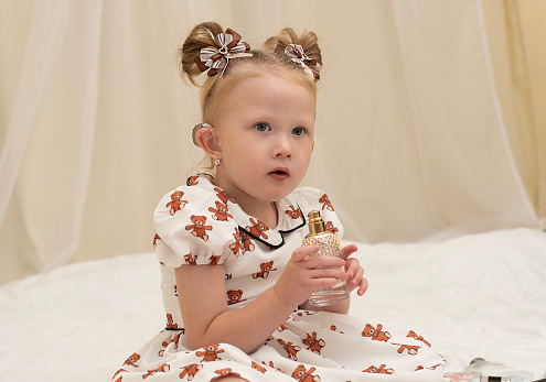 A beautiful little girl wearing a hearing aid sits on the bed in a beautiful dress and plays with her mother's perfume. Sniffs the aromas. Concept. Close-up.
