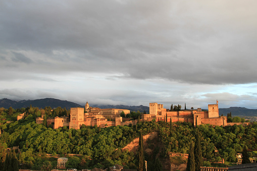 View of Alhambra - Granada in the dusk, with yellowish sunlight and grey cloudy  sky above it.