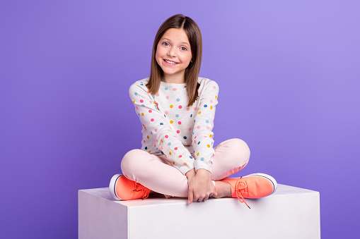 Photo of pretty sweet small girl wear dotted sweater sitting white platform isolated violet color background.