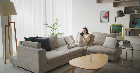 Young South Korean Woman Engaged and Connected Online, Enjoying Leisure Time with Her Smartphone, Spending Time Resting on a Sofa and Exploring Social Media and Entertainment Apps