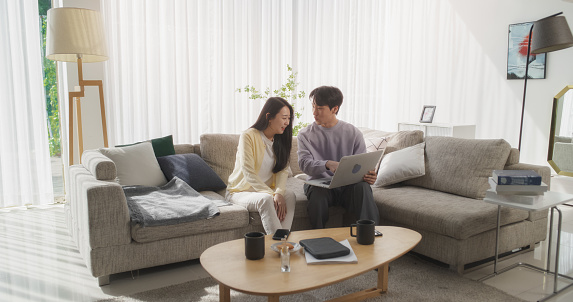 Young and Loving Asian Couple are Browsing Internet, Using Laptop Computer at Home, Planning an Exciting Travel Itinerary for Their Romantic Trip While Sitting on a Couch
