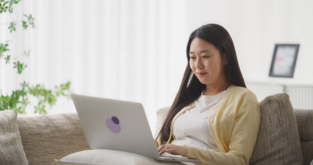 молодая южнокорейская женщина вовлечена и подключена к интернету, наслаждаясь досугом со своим смартфоном, проводя время, отдыхая на диван - проводя поиск стоковые фото и изображения