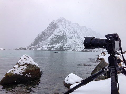 Digital camera on a tripod capturing the snowy winter landscape at the shore of on of the islands of the Lofoten archipel near Heamingsvear in Norway The mountains are covered in snow and there is ice floating on the water.