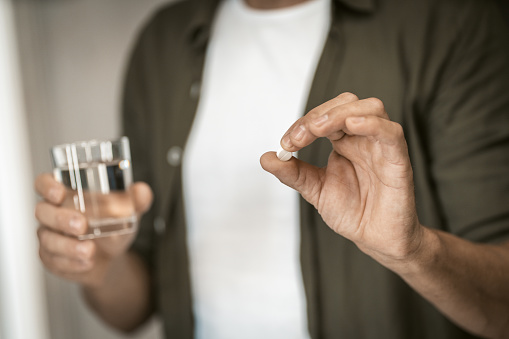 Close-up of man holding pill in one hand and glass of water in other, in comfort of his home. Selective soft focus draws attention to the man's hands, emphasizing the act of taking medicine. The pill and glass of water are in sharp focus, creating a sense of immediacy and importance. High quality photo