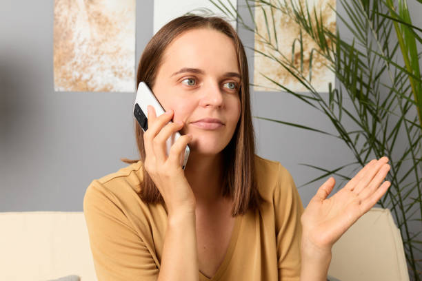 istock Cellular connection. Smartphone call. Dialing friends. Communication gadget. Phone discussion. Puzzled confused dark haired woman wearing beige T-shirt talking via phone at home sitting on couch. 1714853988