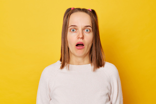 Shocked astonished teenage girl in jumper with ponytails stares bugged eyes feels anxious looks in disbelief hears something impressive isolated over yellow background.