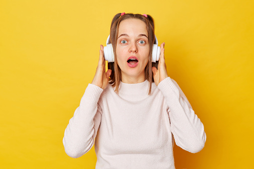 Astonished teen girl wearing jumper and headphones posing isolated over yellow background staring bugged eyes having startled expression being shocked by something listens music via wireless earphones
