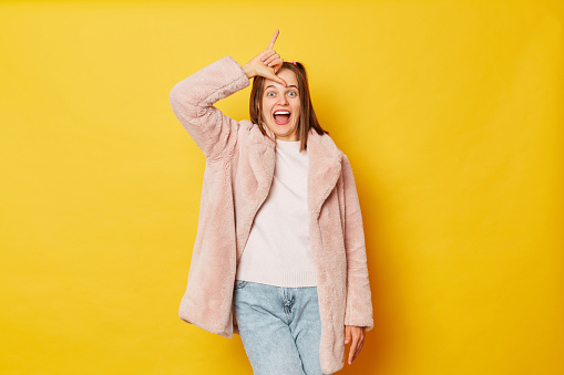 Playful crazy brown haired girl wearing pink fur coat with ponytails isolated over yellow background makes looser gesture over forehead screaming with excitement.