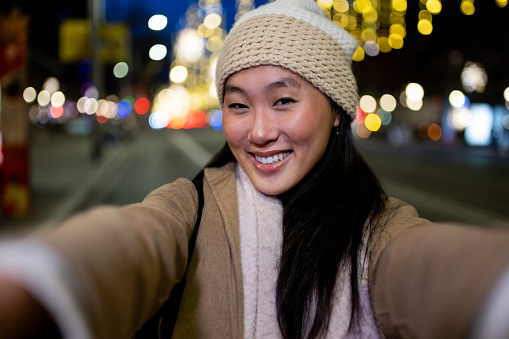 Joyful young woman taking a selfie at night with beautiful christmas lights. Beautiful cheerful female taking a picture in the street wearing a hat.