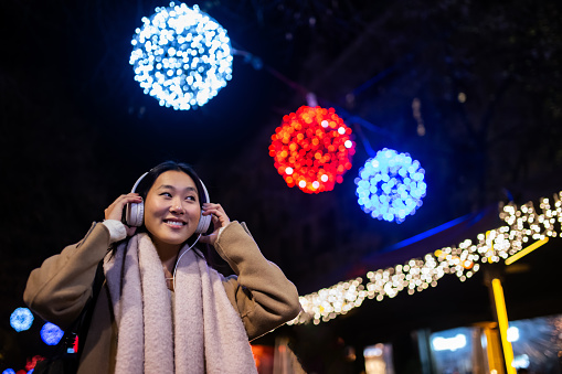Young happy woman wearing headphones with christmas lights behind. Joyful girl listening to music at the street during winter.