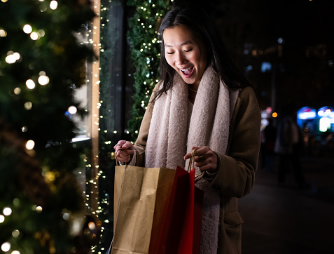 Surprised young lady looking inside shopping bags in the street. Cheerful female holding shopping bags in Christmas at night.