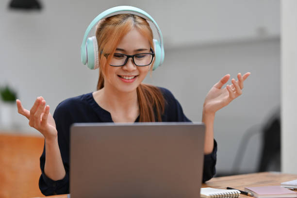 Young millennial woman wearing headphones and gesturing while using laptop for Online class. Young millennial woman wearing headphones and gesturing while using laptop for Online class. online tutors stock pictures, royalty-free photos & images