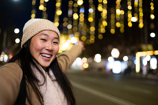 Young cheerful girl taking a selfie in the street at night. Happy woman wearing warm hat looking at camera in winter.